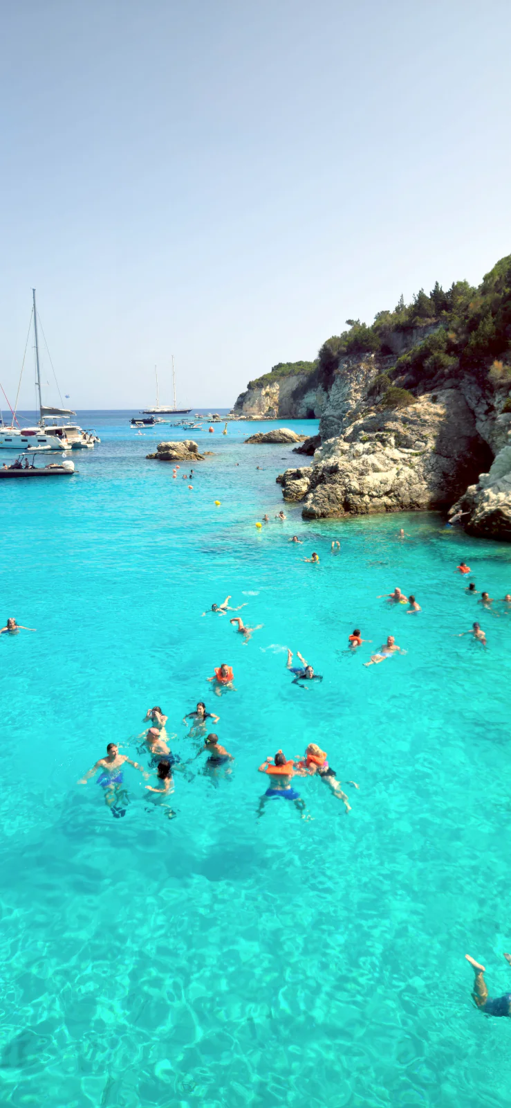 Krystalicznie czysta, turkusowa woda na plaży Voutoumi na Antipaxos, pełna łodzi i pływających ludzi.