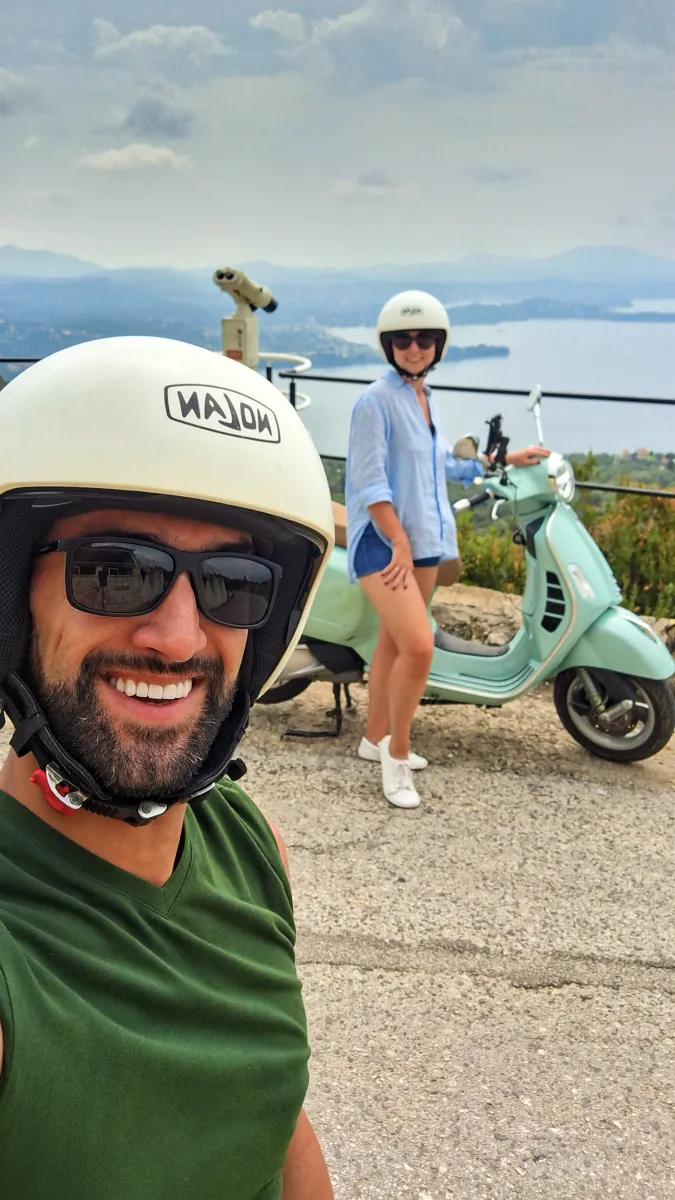 Adam and Joanna from Millennials Do Travel posing with their rented Vespa GTS 300 scooter on a scenic viewpoint in Corfu.