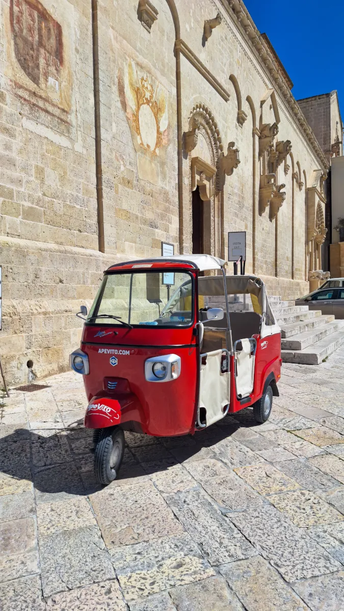 Red Piaggio Ape Calessino three-wheeler parked in front of an ancient stone building in Matera Sassi district.
