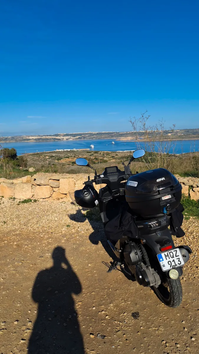 A black rental scooter equipped with a GIVI top case and helmet parked at a scenic viewpoint in Malta, showcasing travel gear setup.