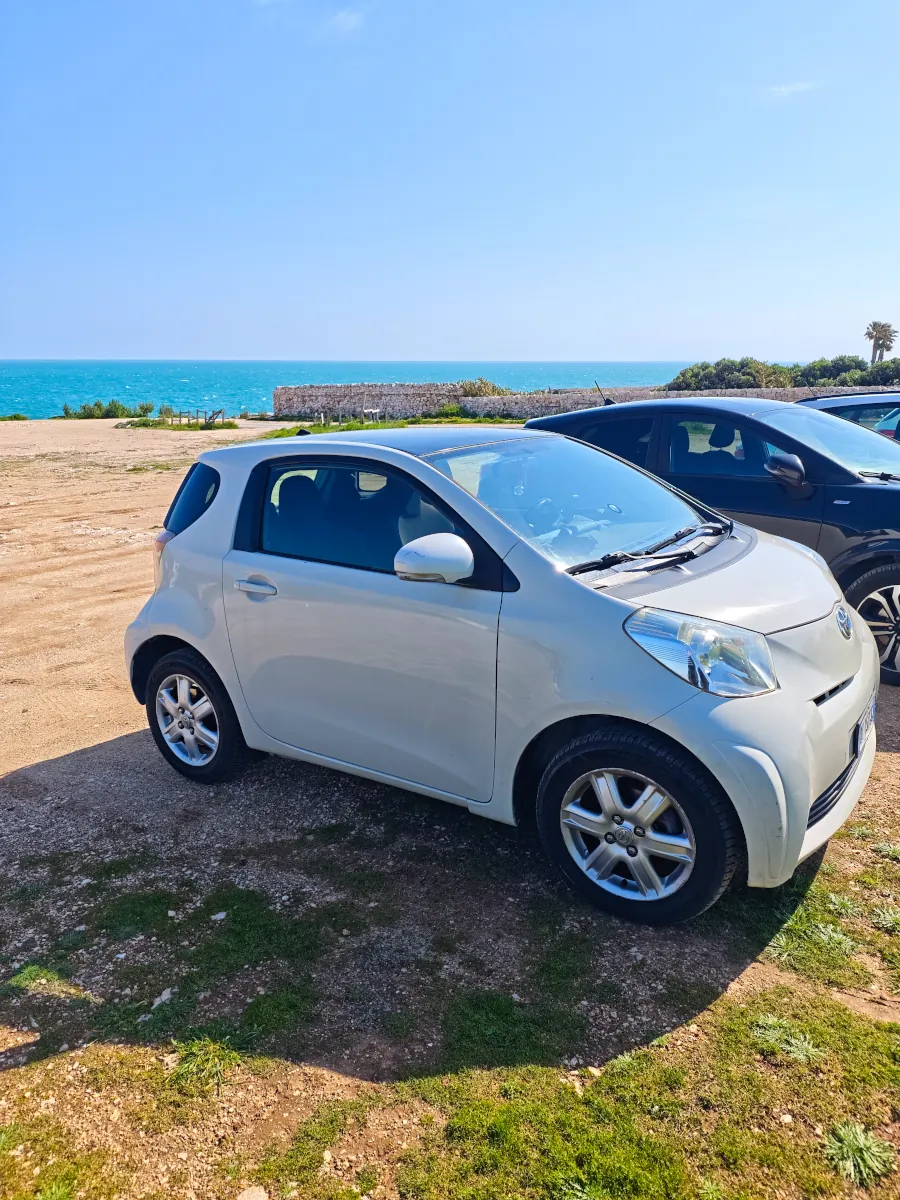 Compact white Toyota iQ parked on a scenic coastal road in Puglia with the blue Adriatic Sea in the background.