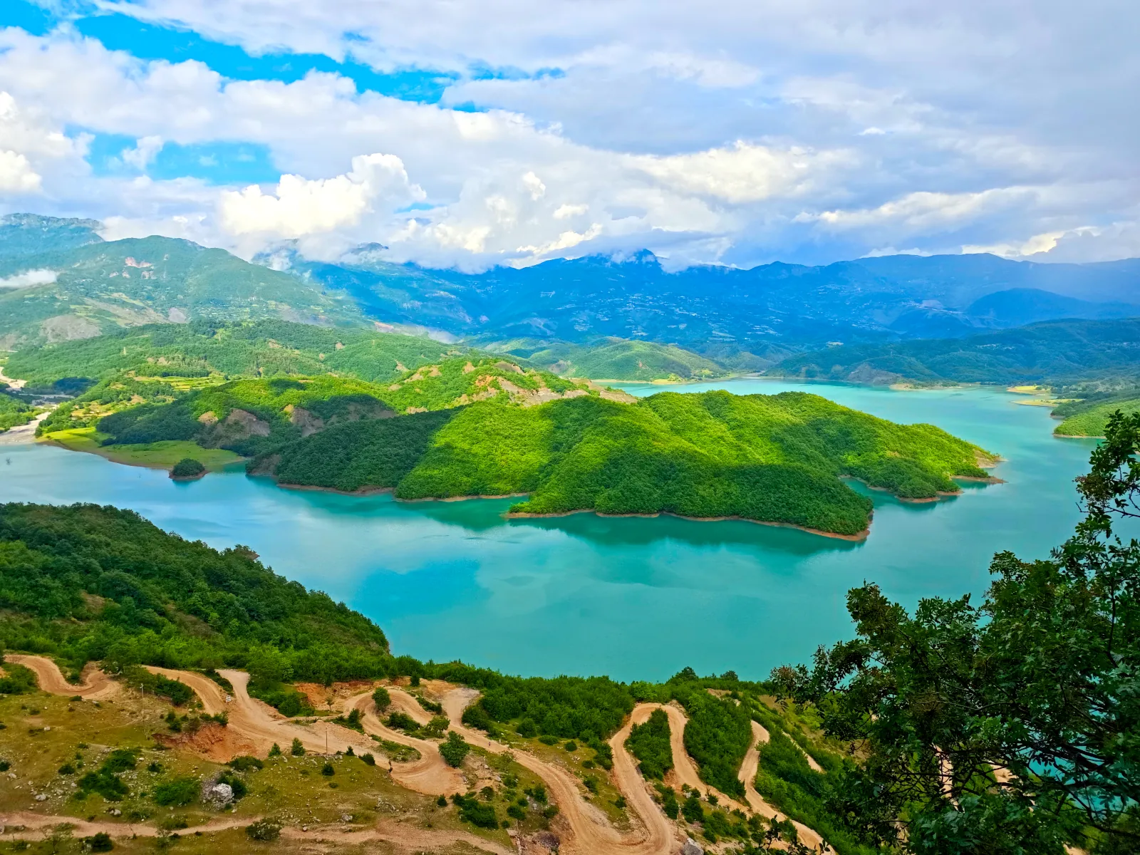 Turquoise waters of Lake Bovilla reservoir surrounded by mountains, with the winding rocky road to the viewpoint.