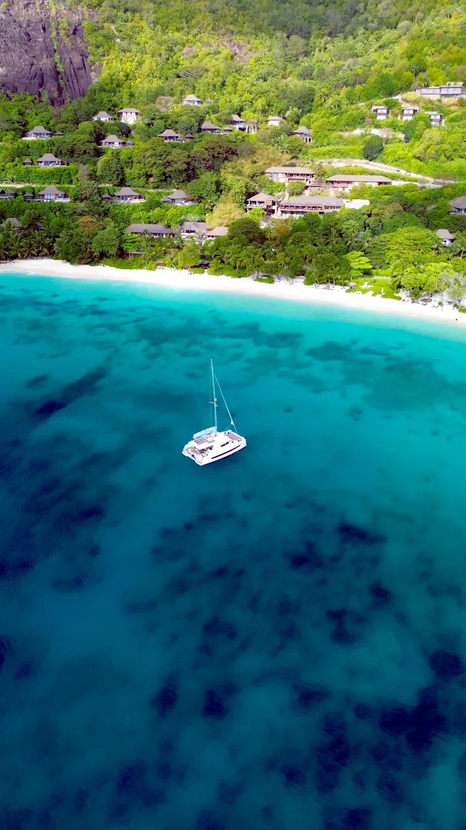 Aerial drone view of a catamaran anchored in the turquoise bay of Petite Anse, Mahé, with Four Seasons luxury villas on the green hillside.