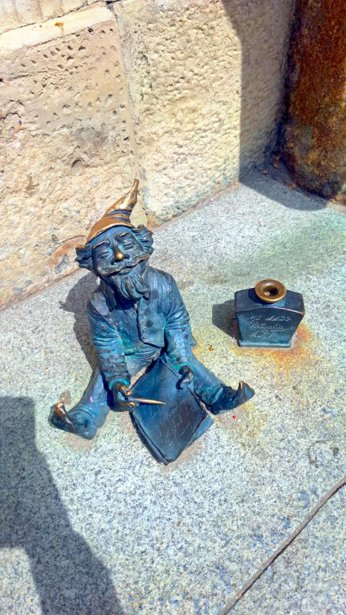 Small bronze Wrocław dwarf figurine sitting on a granite stone step holding a pen next to an inkwell.